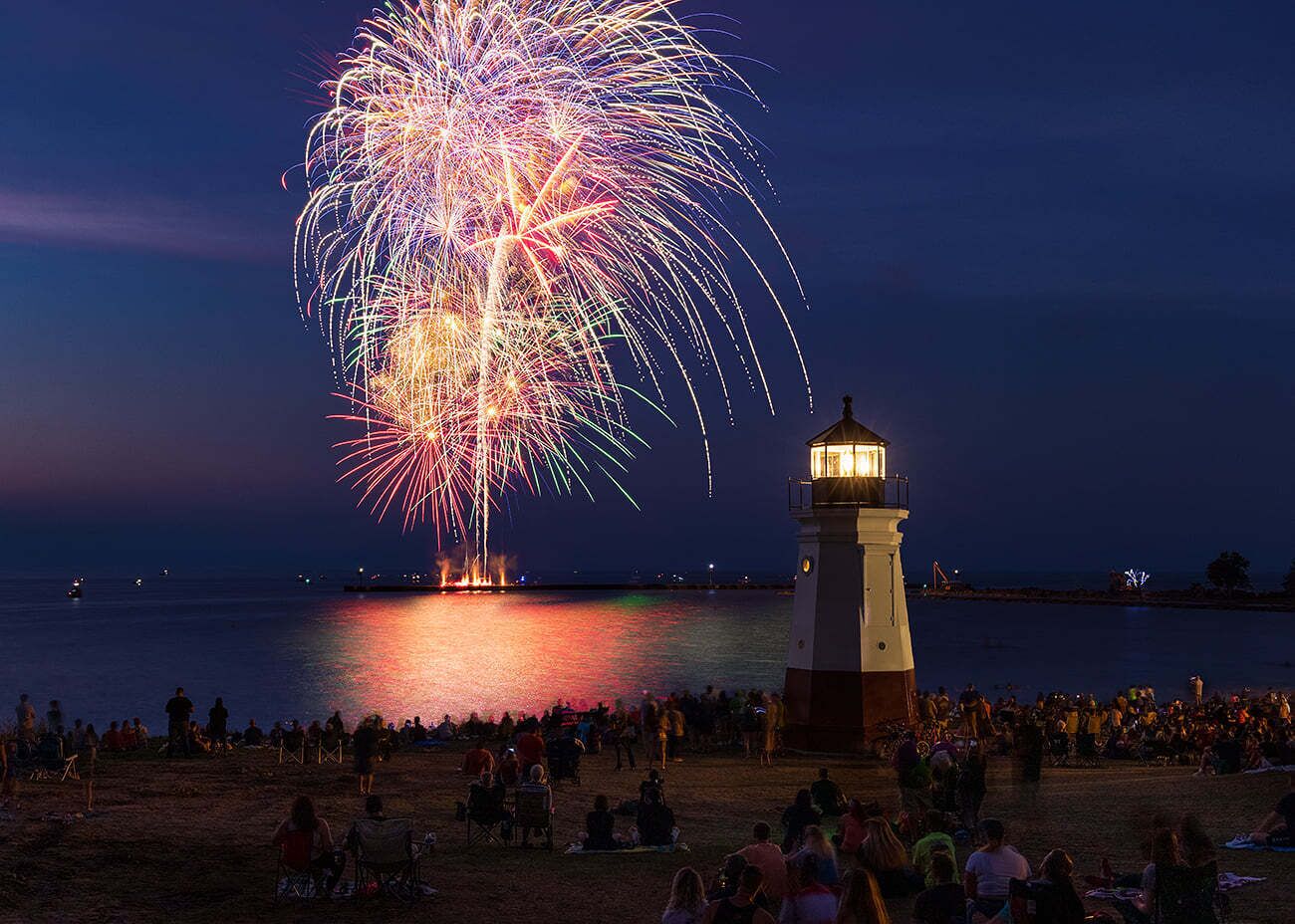 A lighthouse by the sea with a crowd watching fireworks.
