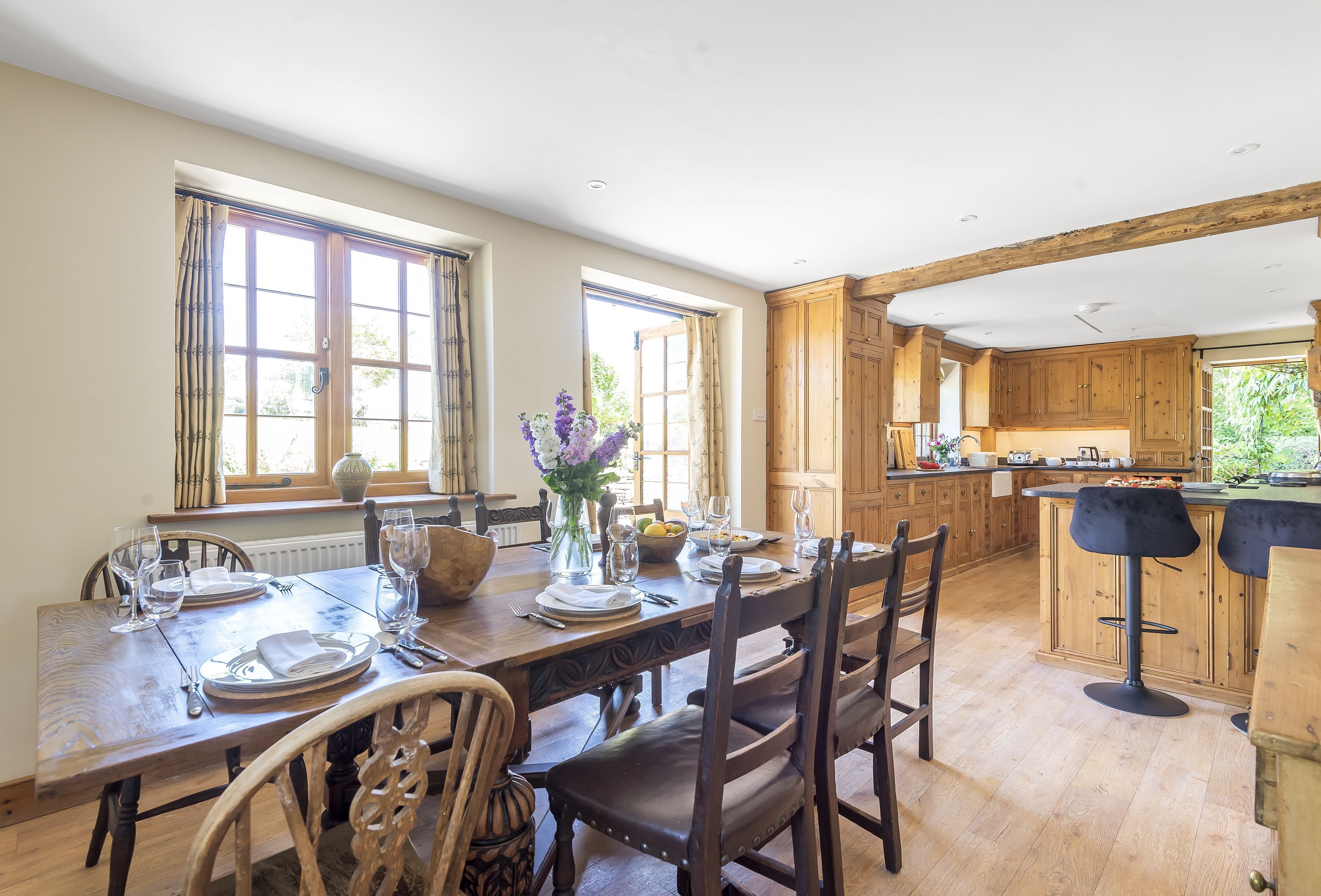 Comedor con una mesa de madera dispuesta para cuatro personas, rodeada de sillas. Se ve una cocina con armarios de madera y una barra en el fondo.