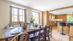 Comedor con una mesa de madera dispuesta para cuatro personas, rodeada de sillas. Se ve una cocina con armarios de madera y una barra en el fondo.
