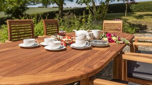 Una mesa de madera dispuesta para un servicio de té, con tazas, platillos y una tetera, rodeada de sillas y un arreglo floral.