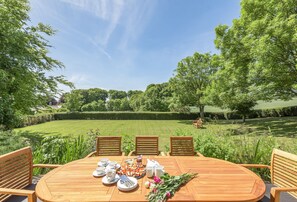 An outdoor dining area with a wooden table set for a meal, surrounded by chairs, and overlooking a well-manicured lawn and trees.