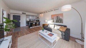 A modern living room with a grey sofa, a white coffee table, and a large floor lamp. There is a kitchen in the background with a dark grey countertop and white cabinets. - Music Row Retreat with Pool and Free Parking, Walk to Bars! (Nashville)