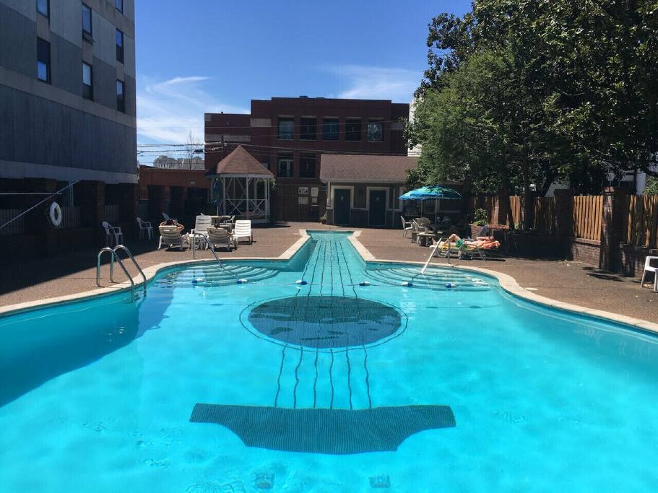 A swimming pool with a central lane, surrounded by lounge chairs and umbrellas, with buildings in the background.
