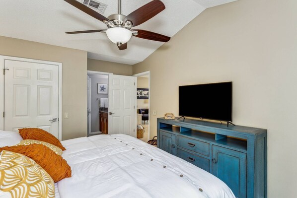 Un dormitorio con una cama, un ventilador de techo, un televisor sobre un aparador azul y una puerta que da a otra habitación.