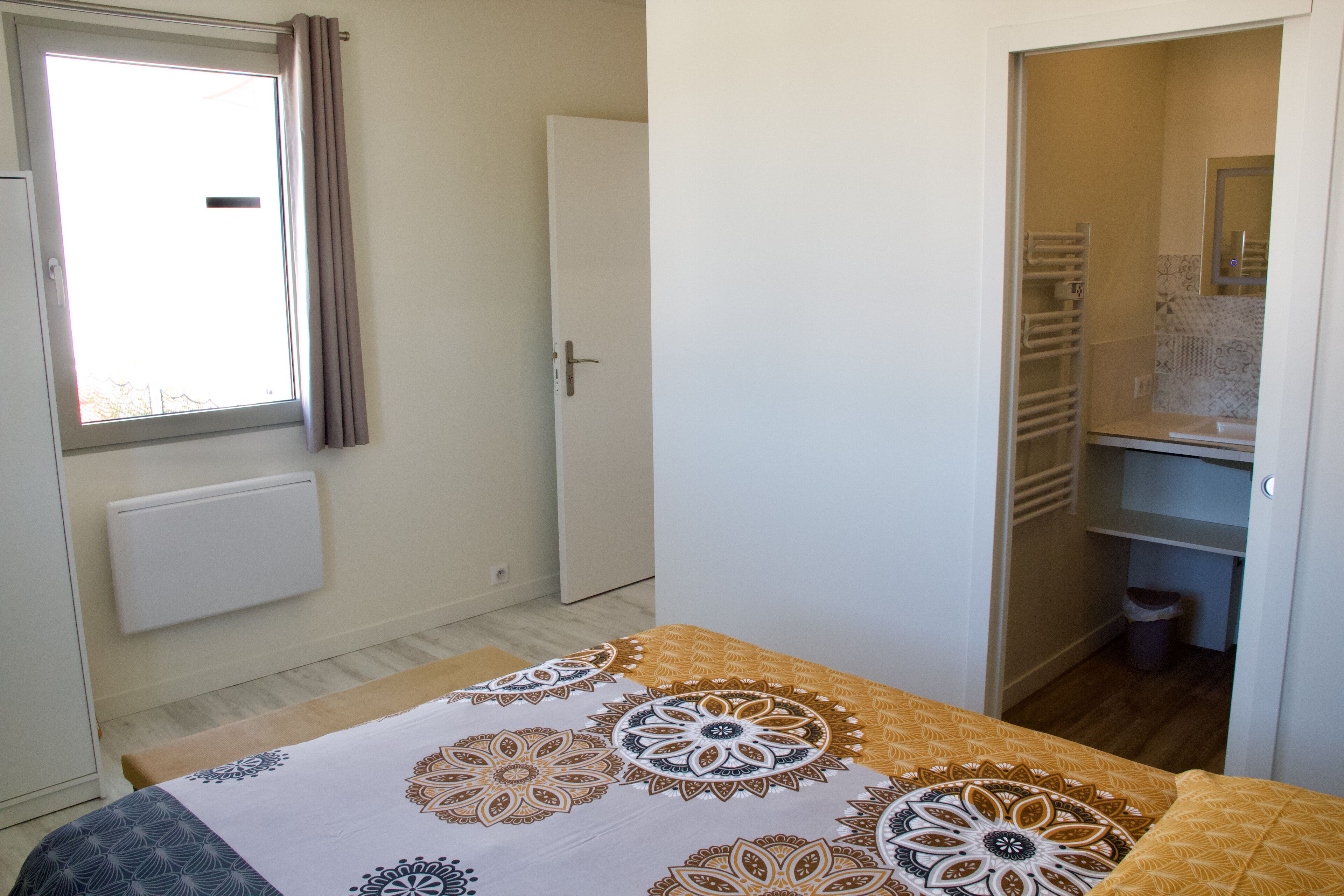 A bedroom with a bed, a window with curtains, a wardrobe, and a bathroom visible through an open door.