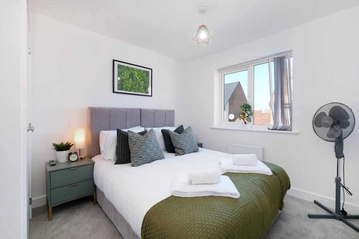 A bedroom with a bed, bedside table, lamp, and a standing fan.