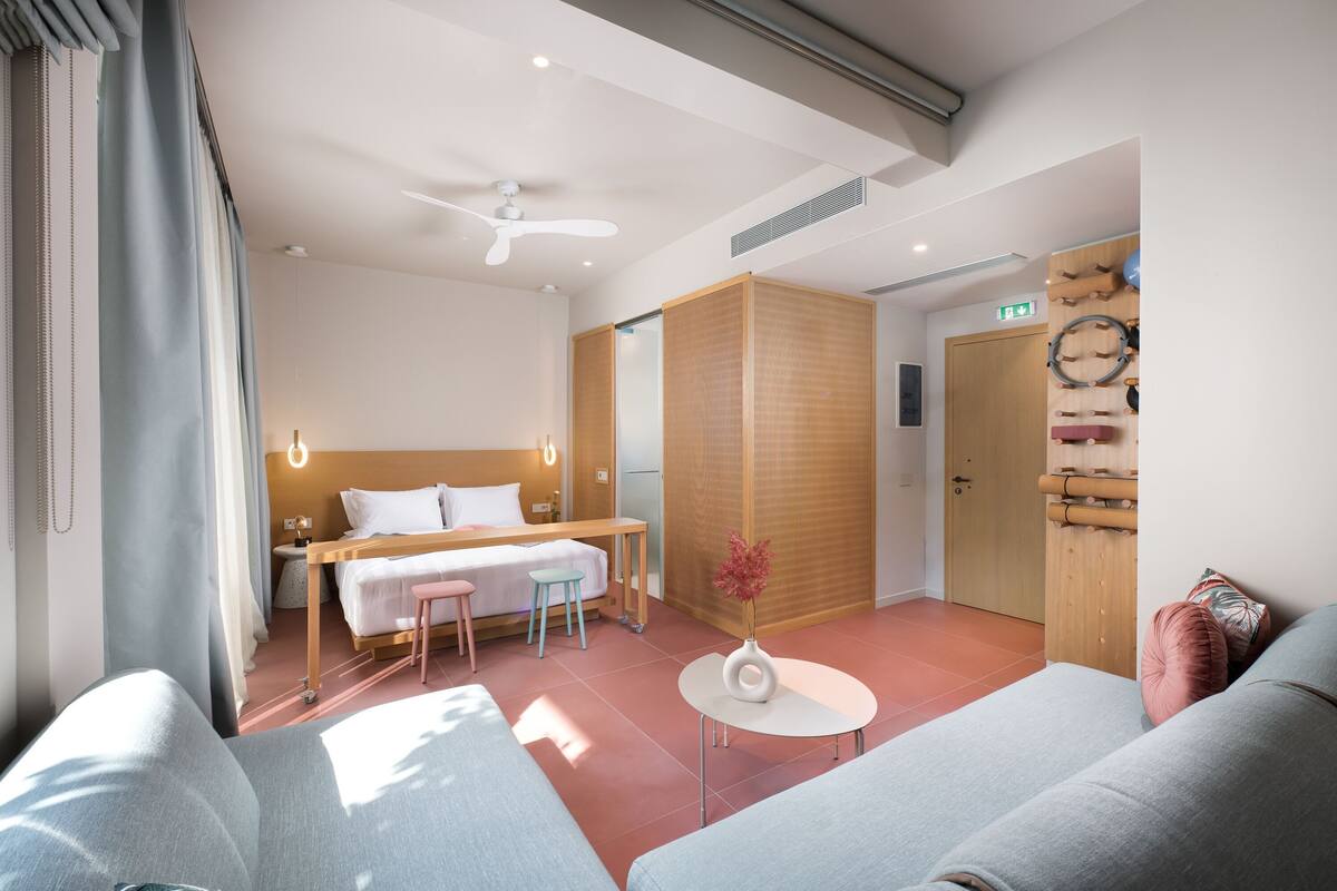 A modern hotel room with a bed, a small table with a vase, a sofa, a ceiling fan, and a wooden partition.
