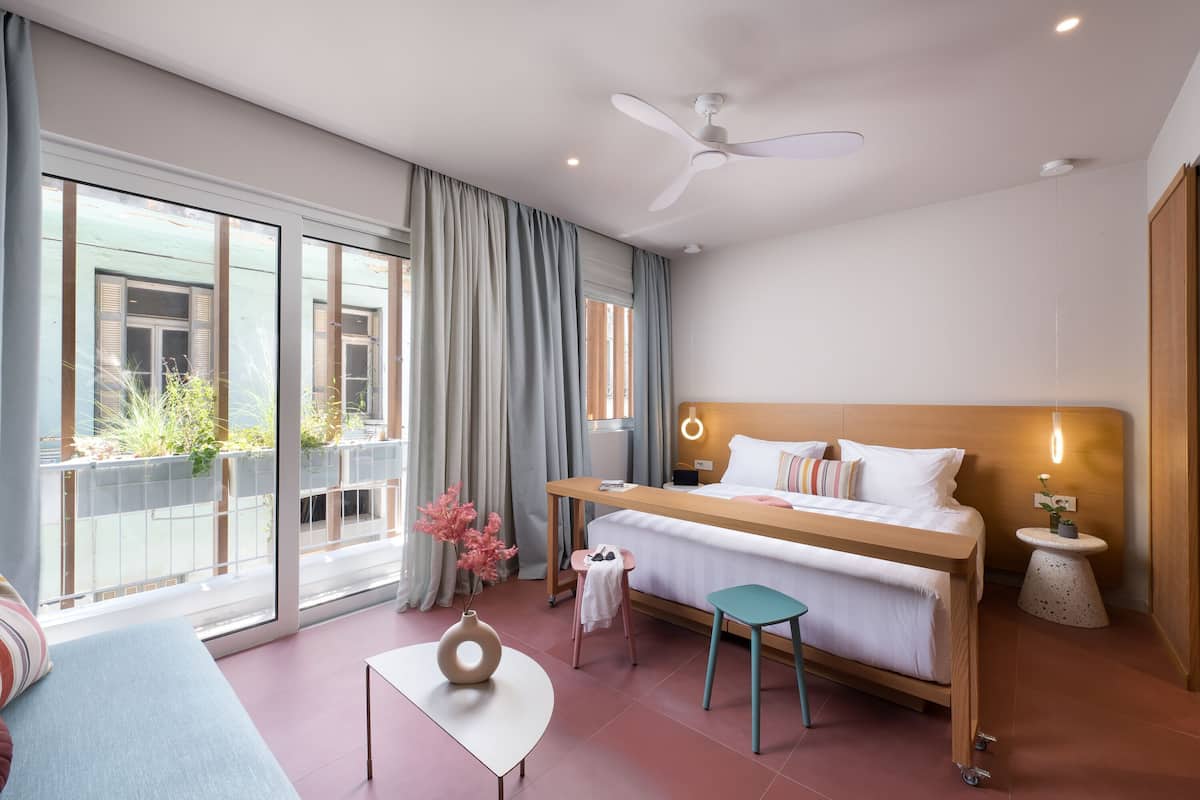 A modern hotel room with a bed, a small table, a stool, a round side table, a ceiling fan, and large windows with curtains.