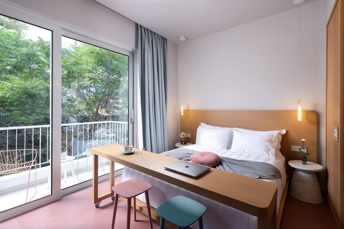 A modern hotel room with a bed, a desk, and a laptop. There is a balcony with a chair and a table.