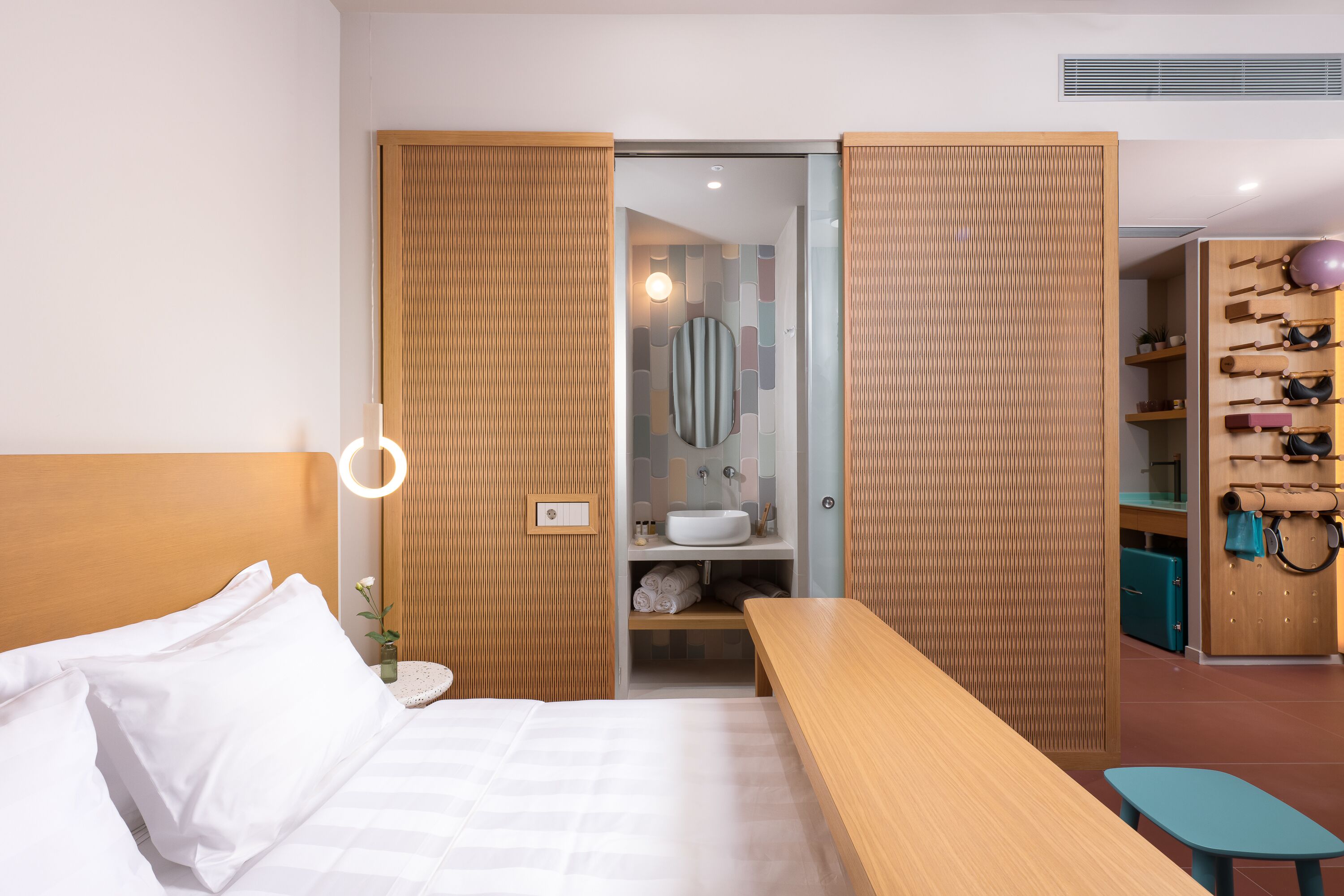 A modern hotel room with a bed, a wooden bench, a small table, a bathroom with a sink and mirror, and a shoe rack with various shoes.