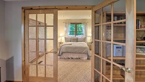 Ein Schlafzimmer mit einer großen Holztür, die ins Badezimmer führt, einem Bett mit grauer Bettdecke und einer Holzbibliothek mit Büchern und Dekorationsobjekten.