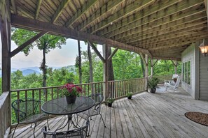 Une terrasse en bois avec une table et des chaises, entourée de plantes en pot et offrant une vue sur une zone boisée avec des montagnes à l’horizon.