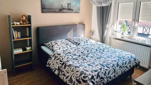 A bedroom with a bed, a bookshelf, a painting, a chandelier, and a window with blinds.