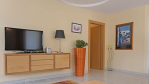 Ein modernes Wohnzimmer mit Flachbildfernseher, einer Lampe, einem Blumentopf, einem Esstisch mit einer Schüssel Snacks und einem Bild an der Wand.