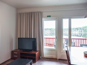 Een kamer met een televisie op een houten kast, een glazen schuifdeur die leidt naar een buitenruimte met een rode balustrade, en een bord boven de deur met de tekst 'VARATIE'.