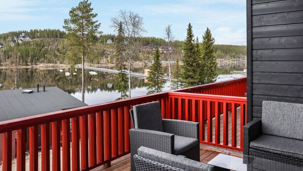 Una terrazza con mobili in vimini che si affaccia su un lago, circondata da alberi e case sullo sfondo.