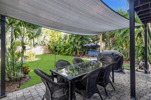 An outdoor patio with a glass-top table and wicker chairs, surrounded by lush greenery and a covered barbecue area.