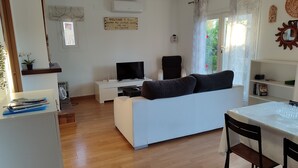A modern living room with a white sofa, a TV, a dining table with chairs, and a kitchen area with a fridge and a microwave.