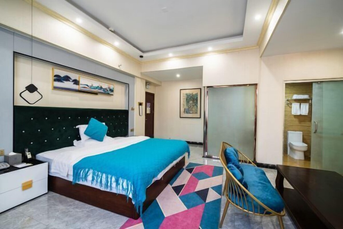 A modern hotel room with a large bed, a bedside table, a chair, and a bathroom visible through a glass door.