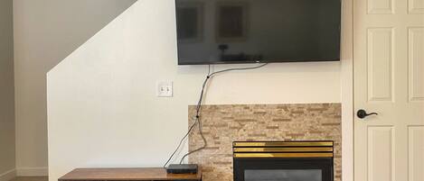 A living room with a flat-screen TV mounted on the wall, a fireplace with a gold frame, and a black entertainment unit with drawers.