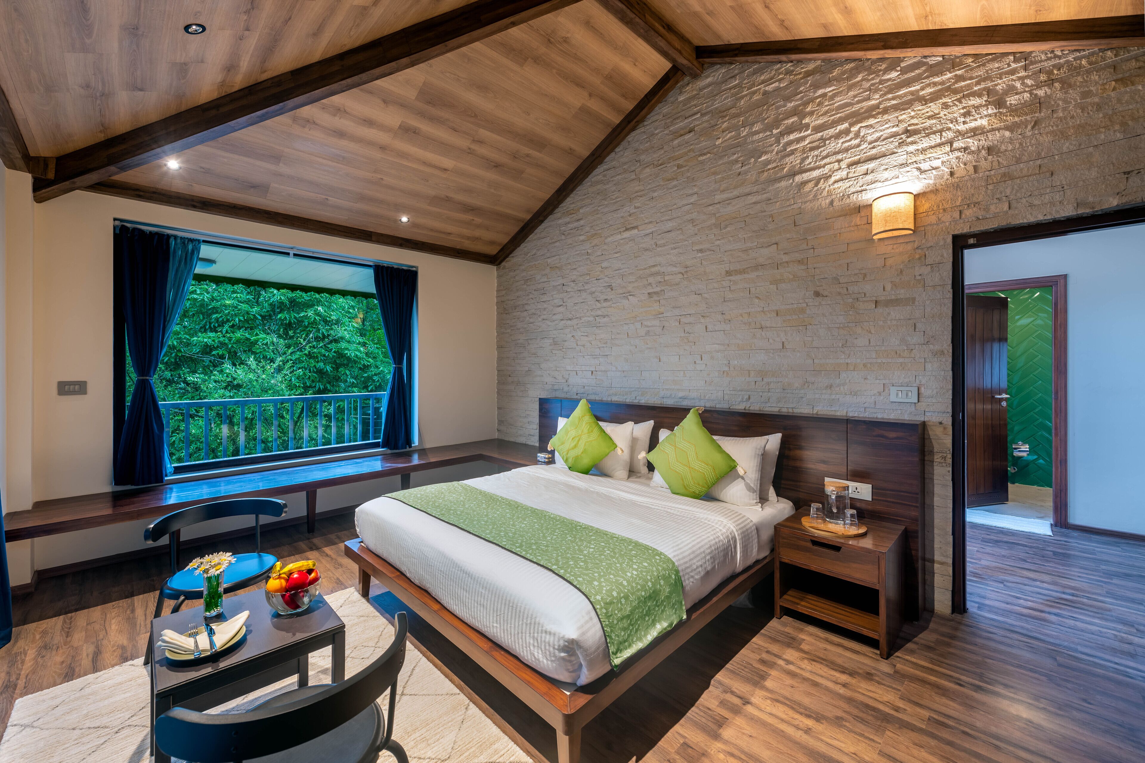 A bedroom with a large bed, a nightstand, a chair, a table with a bowl of fruit, and a window with a view of greenery.