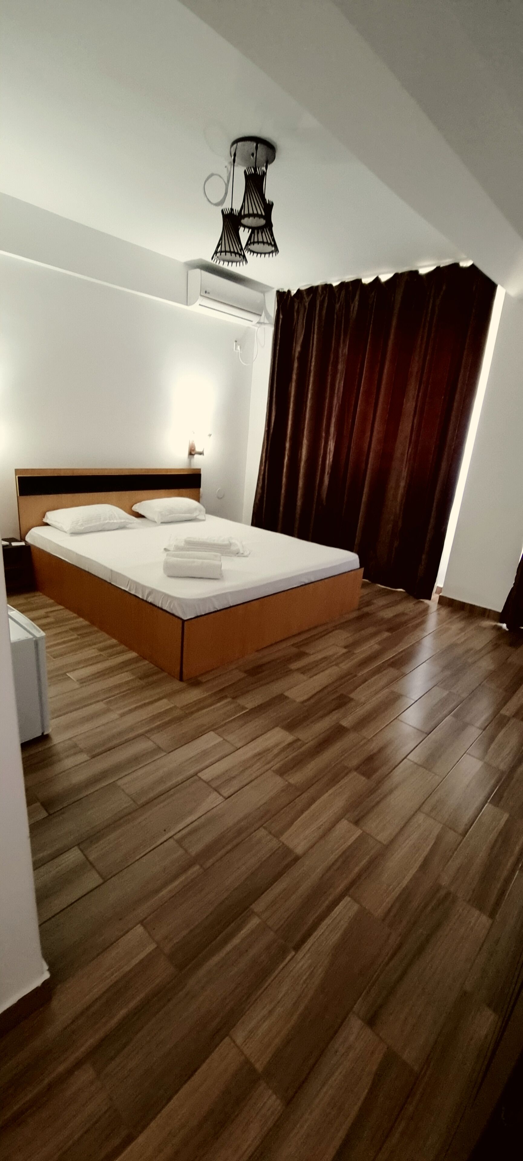 A bedroom with a wooden floor, a bed with white linens, a bedside table, a wall-mounted light fixture, and a window with curtains.