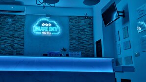 Zona de recepción de un hotel con un letrero de neón que dice 'BLUE SKY HOTEL', un mostrador de recepción y una televisión instalada en la pared.