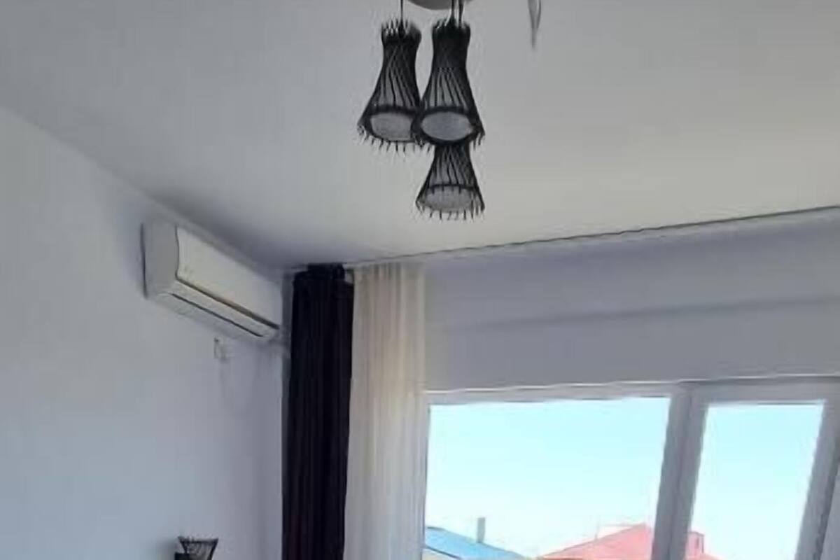 A bedroom with a bed, wooden flooring, a ceiling light fixture, and a view through a window.