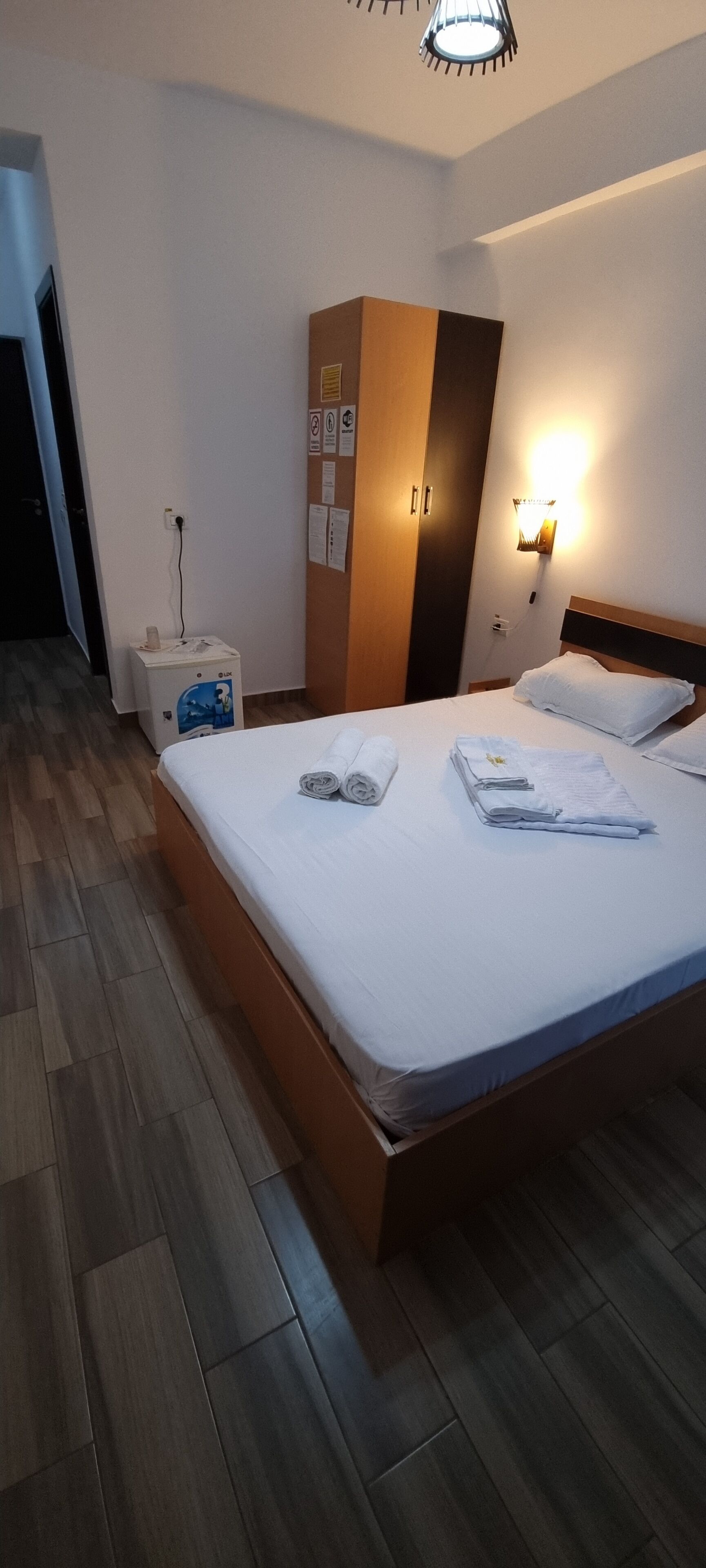 A hotel room with a wooden bed, a wardrobe, a wall-mounted light, and a water dispenser.