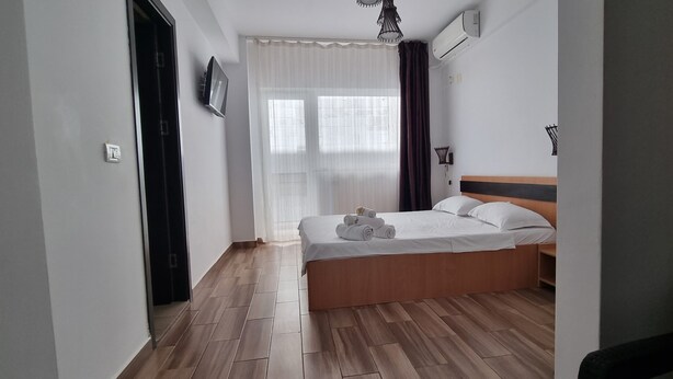 Un dormitorio con cama de madera, ropa de cama blanca, un televisor en la pared y una ventana con cortinas.