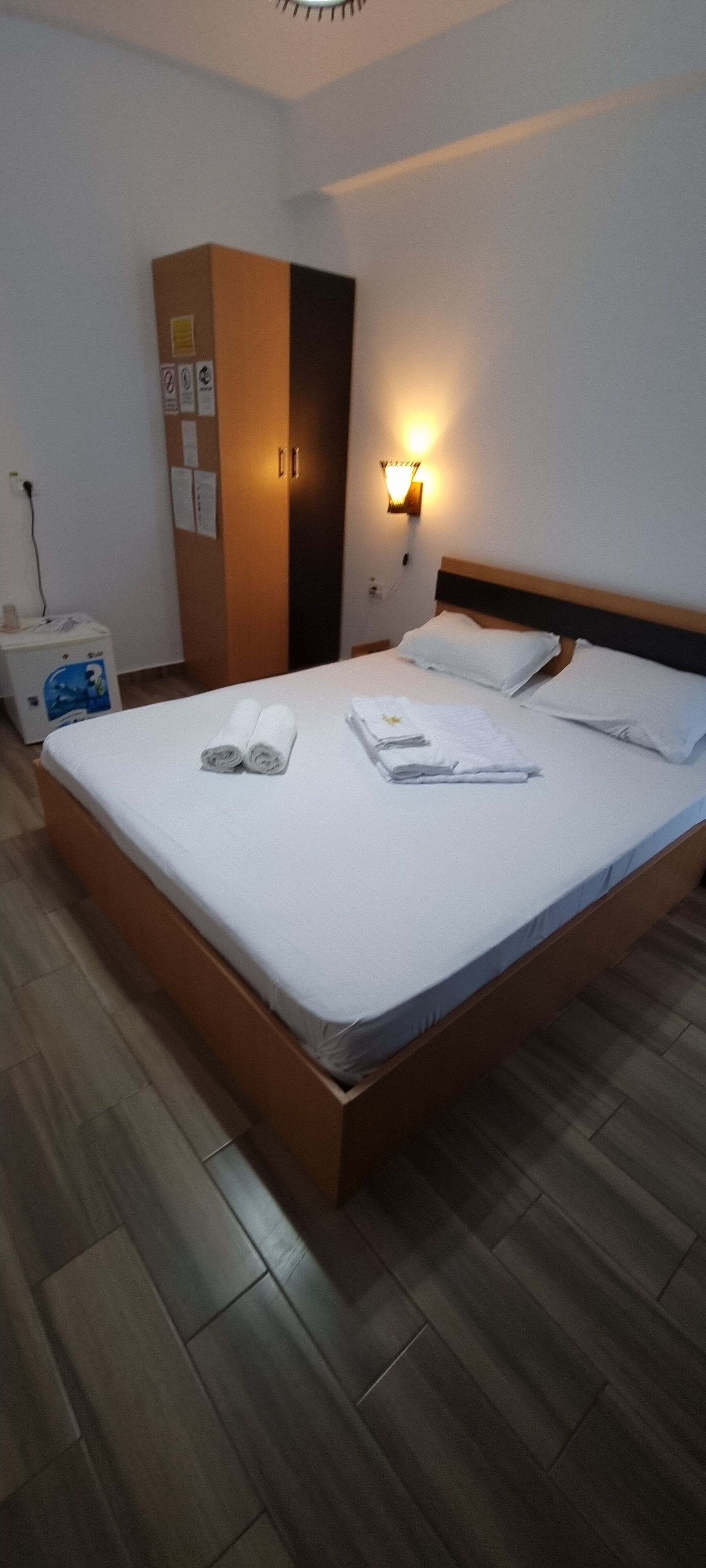 A hotel room with a wooden bed, white linens, a nightstand with a lamp, and a wardrobe.