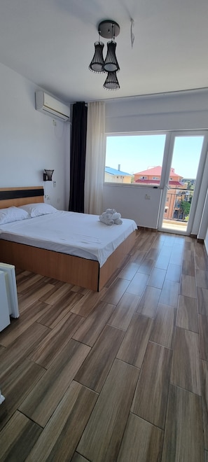 Quarto de hotel com piso de madeira, cama com roupa branca, luminária no teto e vista através de uma janela grande.