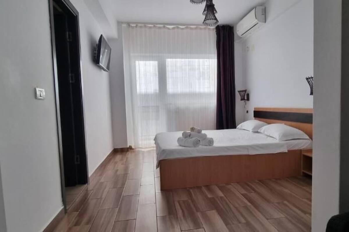 A bedroom with a wooden bed, a window with sheer curtains, a wall-mounted air conditioner, and a door.