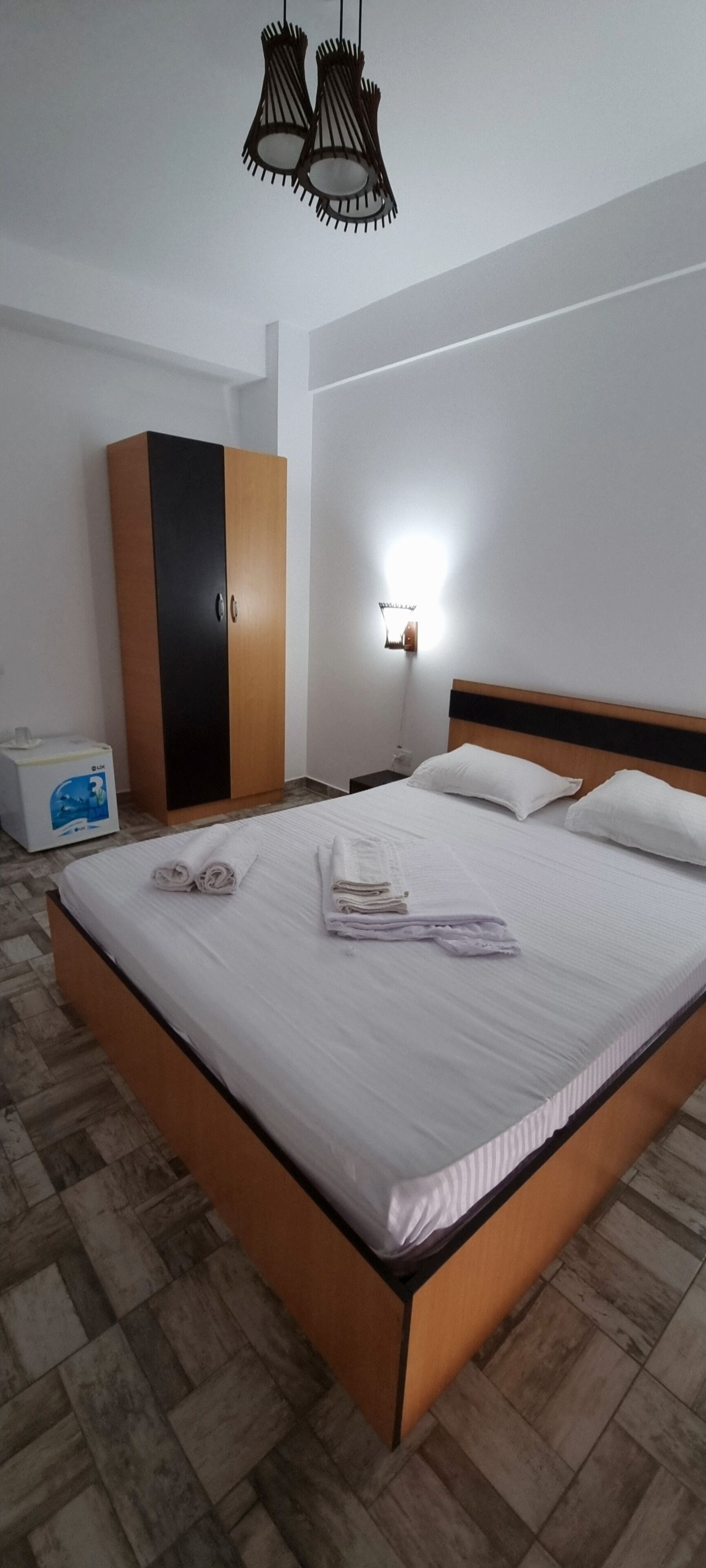 A neatly made bed with rolled towels, a wardrobe, a bedside table with a lamp, and a patterned floor.