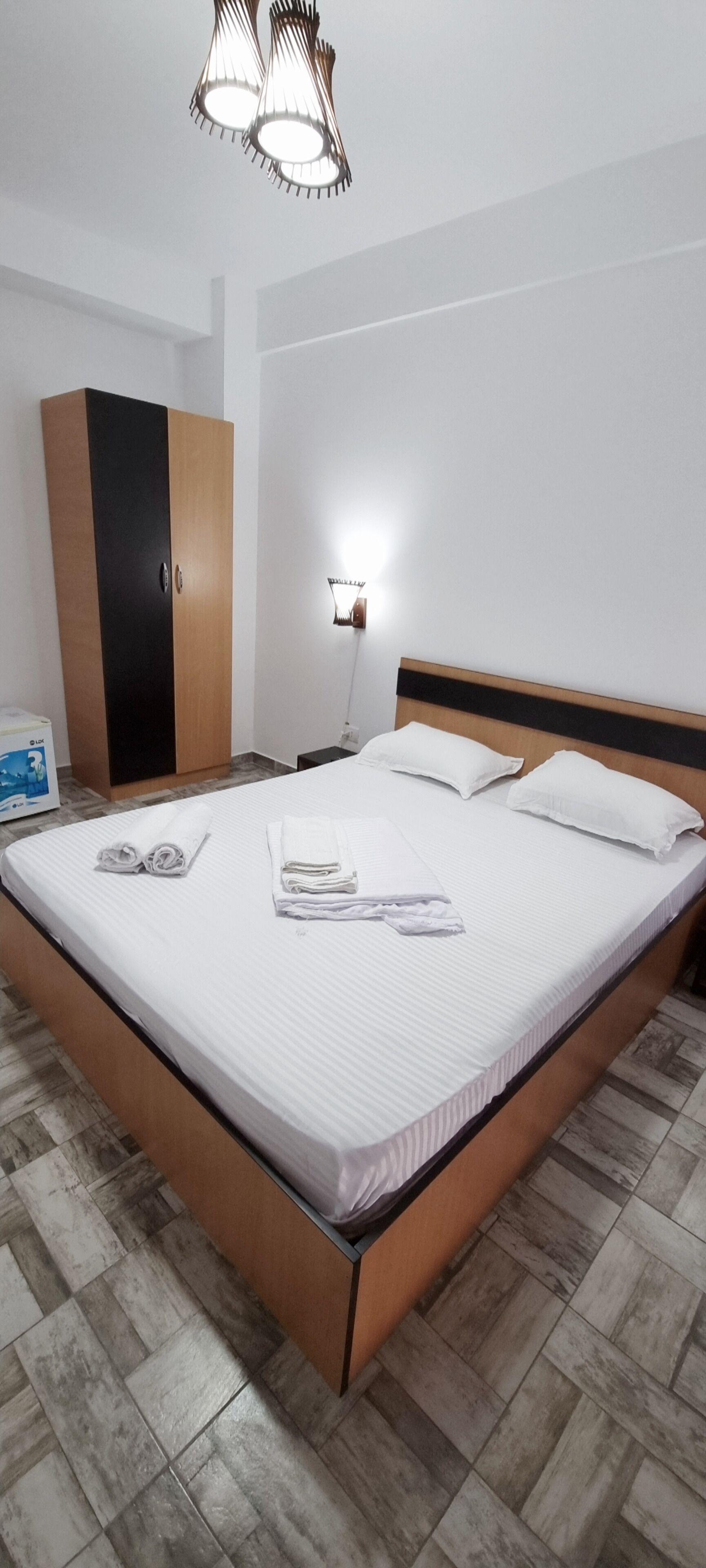 Una cama bien hecha con sábanas blancas y toallas enrolladas, un armario de madera y una mesita de noche con una lámpara.