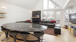 A modern living room with a round marble table, black bar stools, a grey sofa, a fireplace, a TV, and large windows. - Fifth Avenue Unit 5 | Modern & Stylish Condo with A/C and Access to Pool & Hot Tub (Aspen)