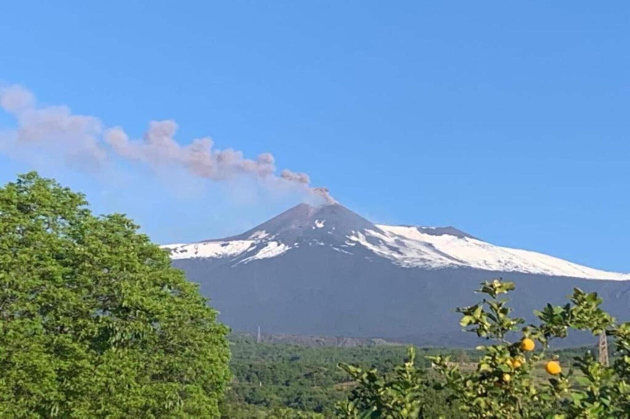 A snow-capped mountain with a visible smoke plume, surrounded by greenery and trees.