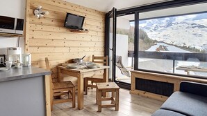 A cozy room with a wooden wall, a dining table with chairs, a TV mounted on the wall, and a large window offering a mountain view. - Renovated studio, center, balcony, 3 pers., 5th floor (Les Menuires)