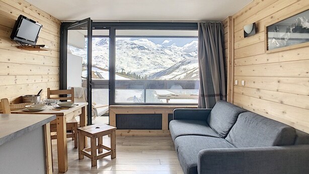 A cozy room with a wooden interior, a gray sofa, a dining table with chairs, and a large window offering a mountain view. - Renovated studio, center, balcony, 3 pers., 5th floor (Les Menuires)