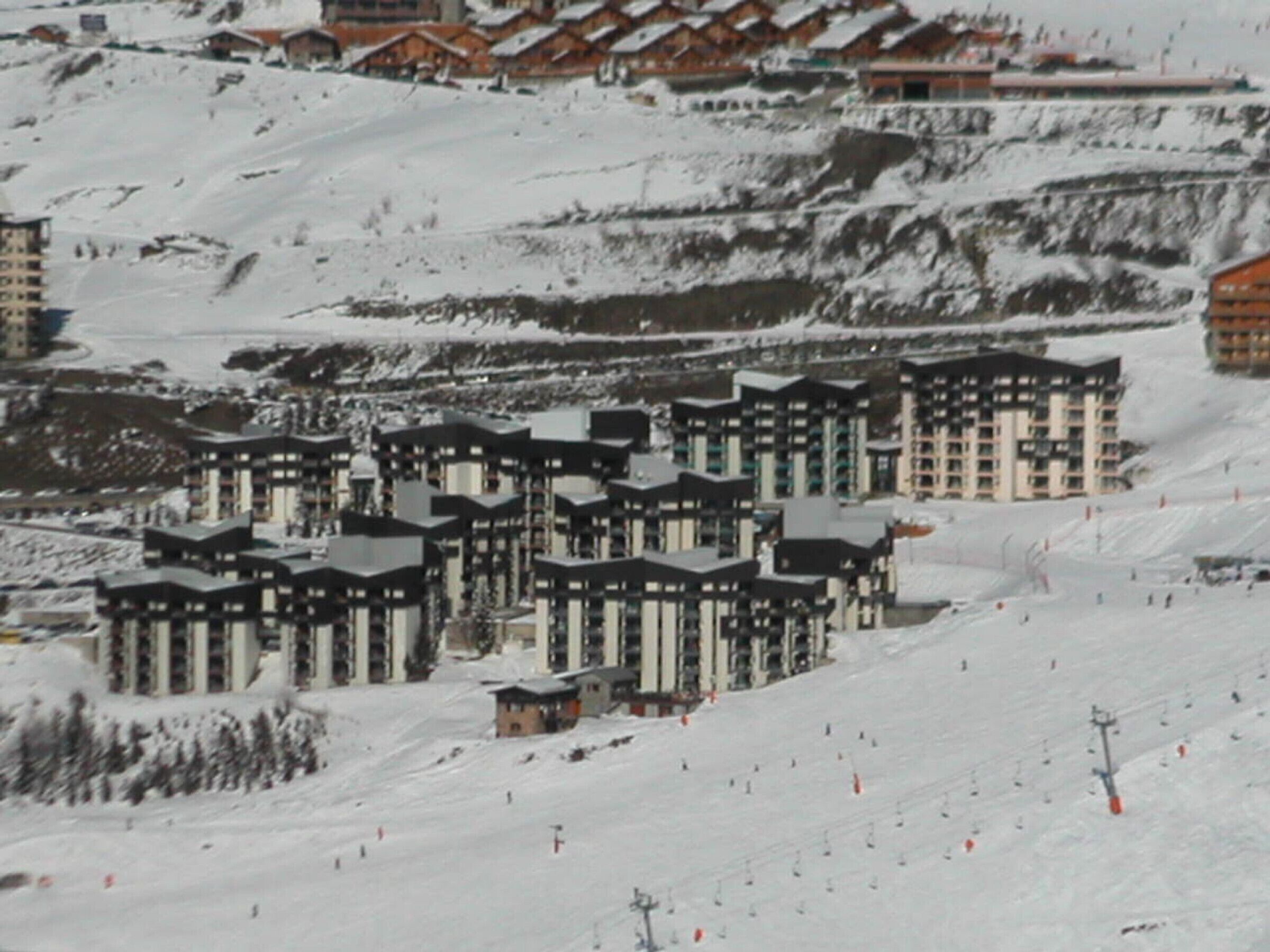 A cluster of modern ski resort buildings with multiple floors and balconies, surrounded by a snowy landscape and ski slopes.