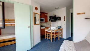 A compact room with a bunk bed, a small kitchenette, a dining table with chairs, and a single bed with a pillow.