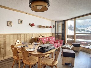 Una habitación acogedora con una mesa de madera, sillas, un sofá con cojines estampados y un ventanal con vistas a un paisaje nevado.