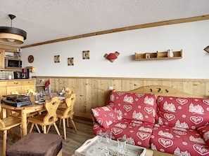 Um quarto aconchegante com uma mesa de jantar de madeira, cadeiras, um sofá com almofadas estampadas em formato de coração, uma cabeceira de madeira e uma cozinha compacta com micro-ondas e cafeteira.