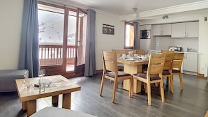 A dining area with a wooden table and chairs, a kitchen with white cabinets, and a large window with curtains. - Comfort & Sauna, Hammam, WIFI, 8 pers, Near slopes (Les Menuires)