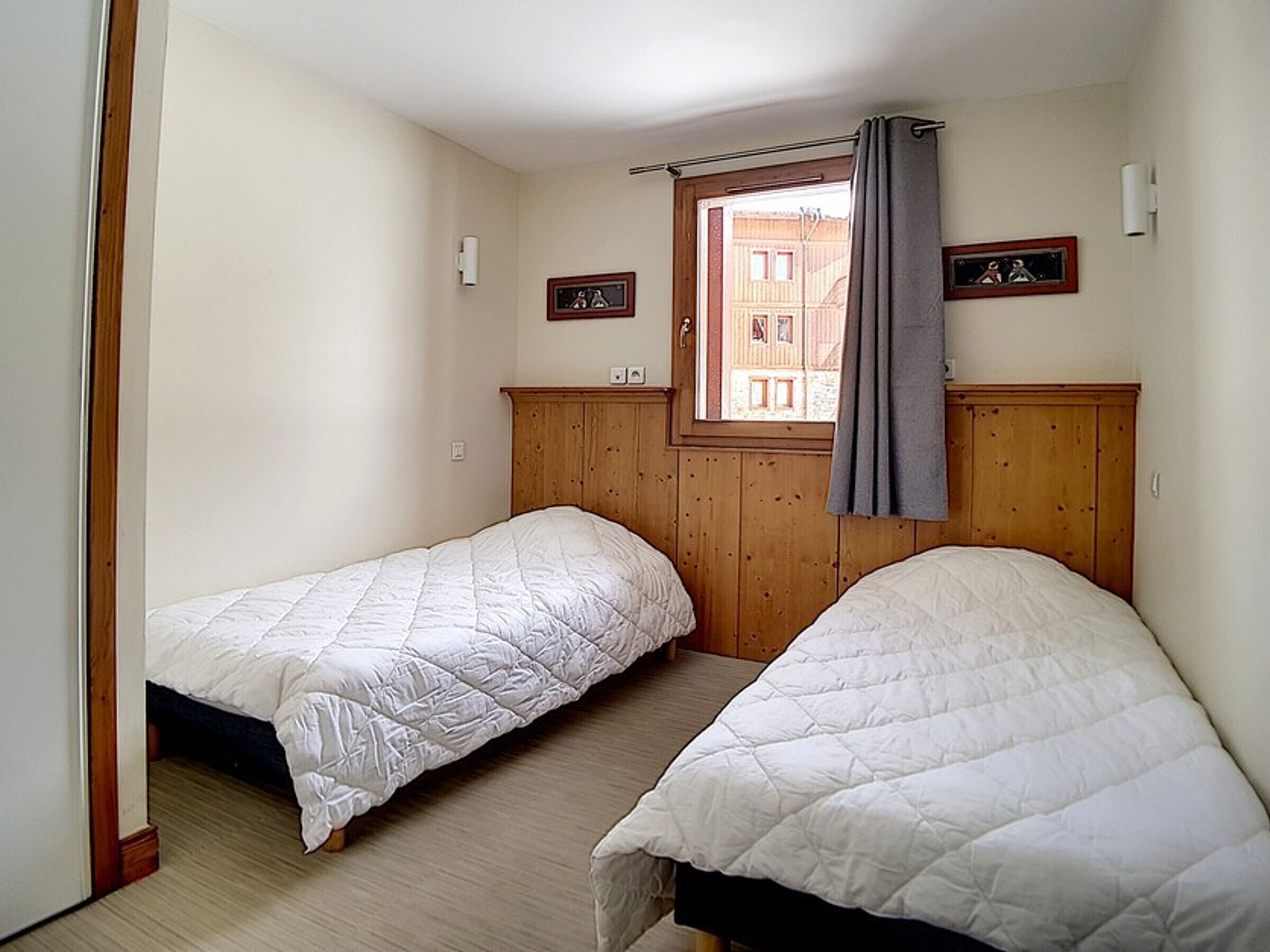 A room with two single beds, wooden headboards, a window with a curtain, and a view of buildings outside.