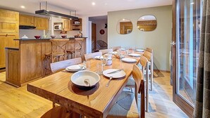 A dining area with a wooden table set for six, a kitchen with wooden cabinets, and a large window with curtains. - Spacious apartment for 10, balcony, parking, near slopes (Saint-Martin-de-Belleville)