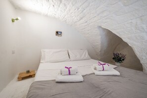 A cozy, white-walled room with a bed, pillows, and neatly folded towels with purple ribbons.