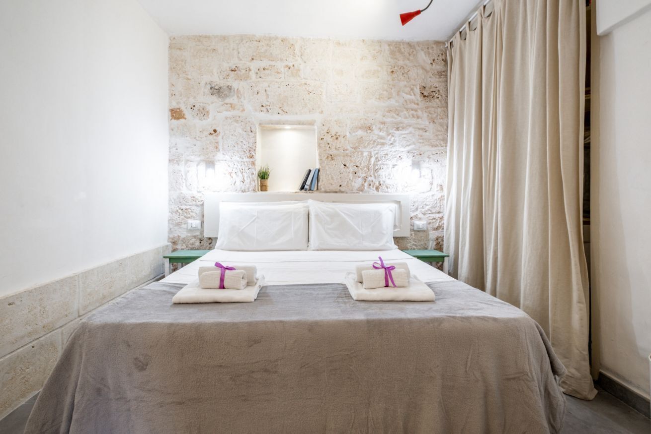 Habitación de hotel con pared de piedra, cama con ropa blanca, una mesita de noche pequeña y ventana con cortinas.