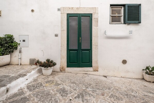 A white building with a green door, a window with shutters, and potted plants.
