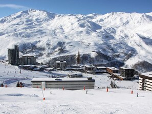 A snow-covered ski resort with multiple buildings, including a prominent church, and a ski slope with skiers. - Studio at the slopes with balcony (Les Menuires)